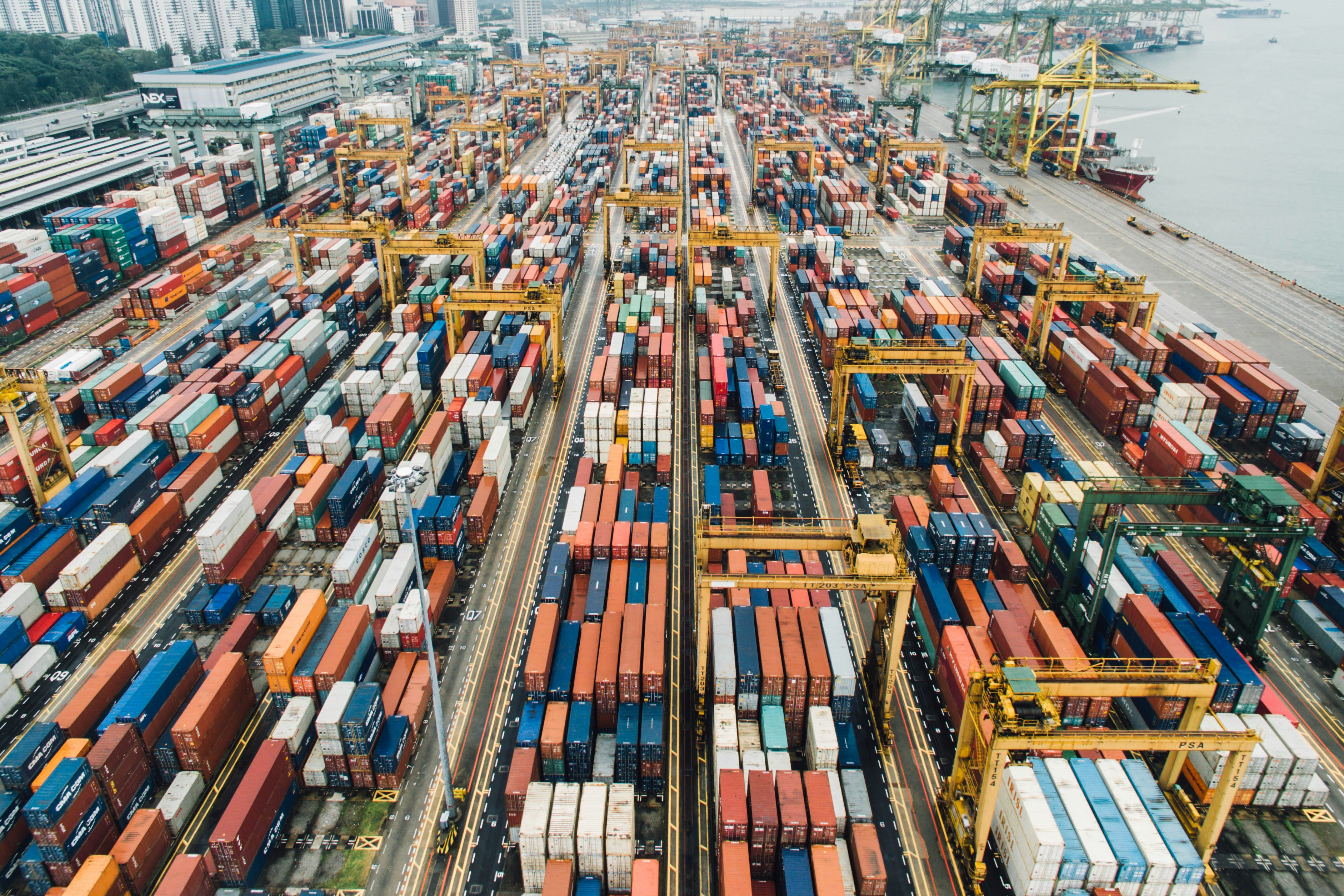 Vista aérea de contêineres organizados em um terminal portuário, com guindastes e navios ao fundo.